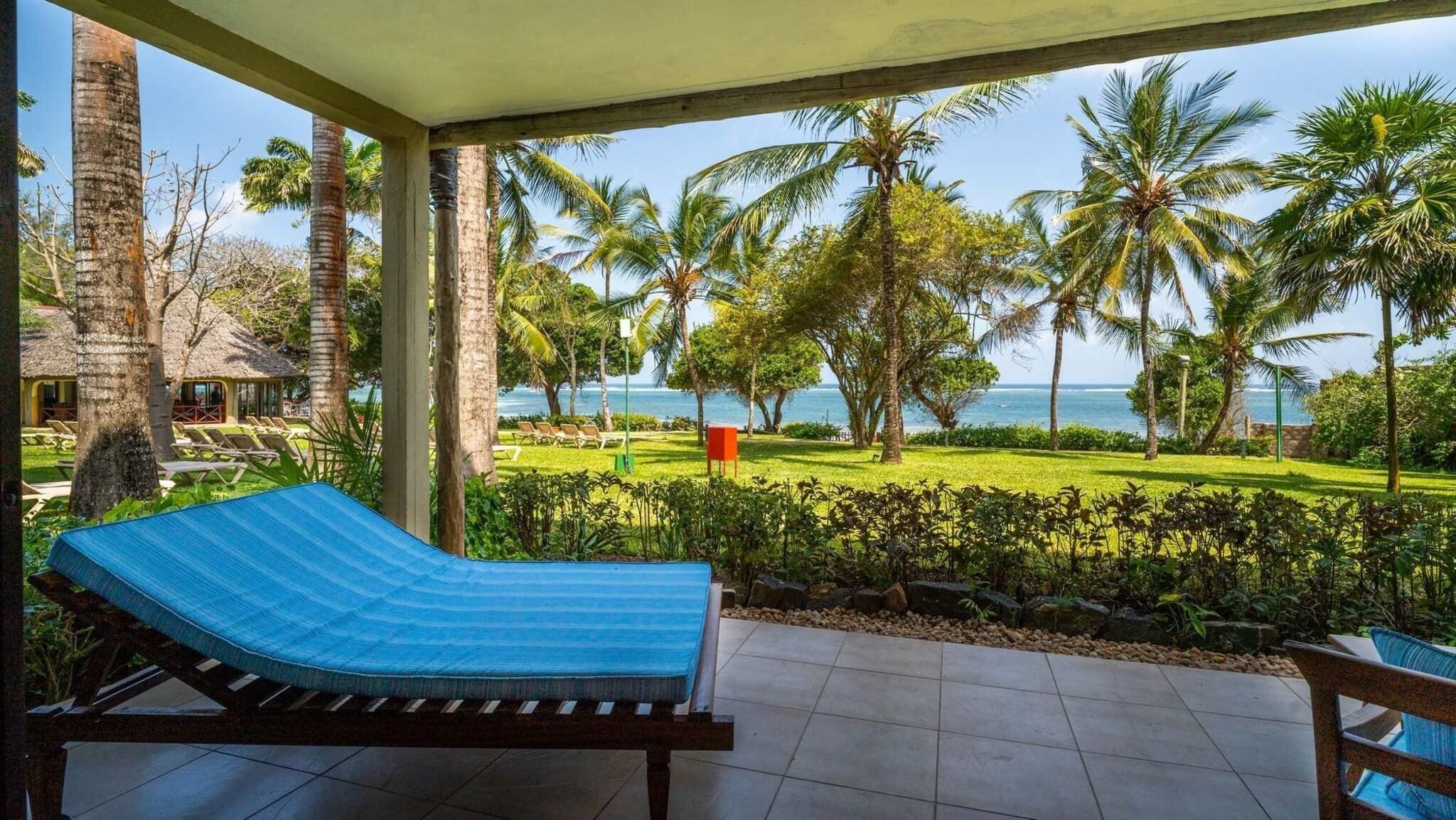 A covered outdoor area with a lounge chair, palm trees, and a view of the ocean.