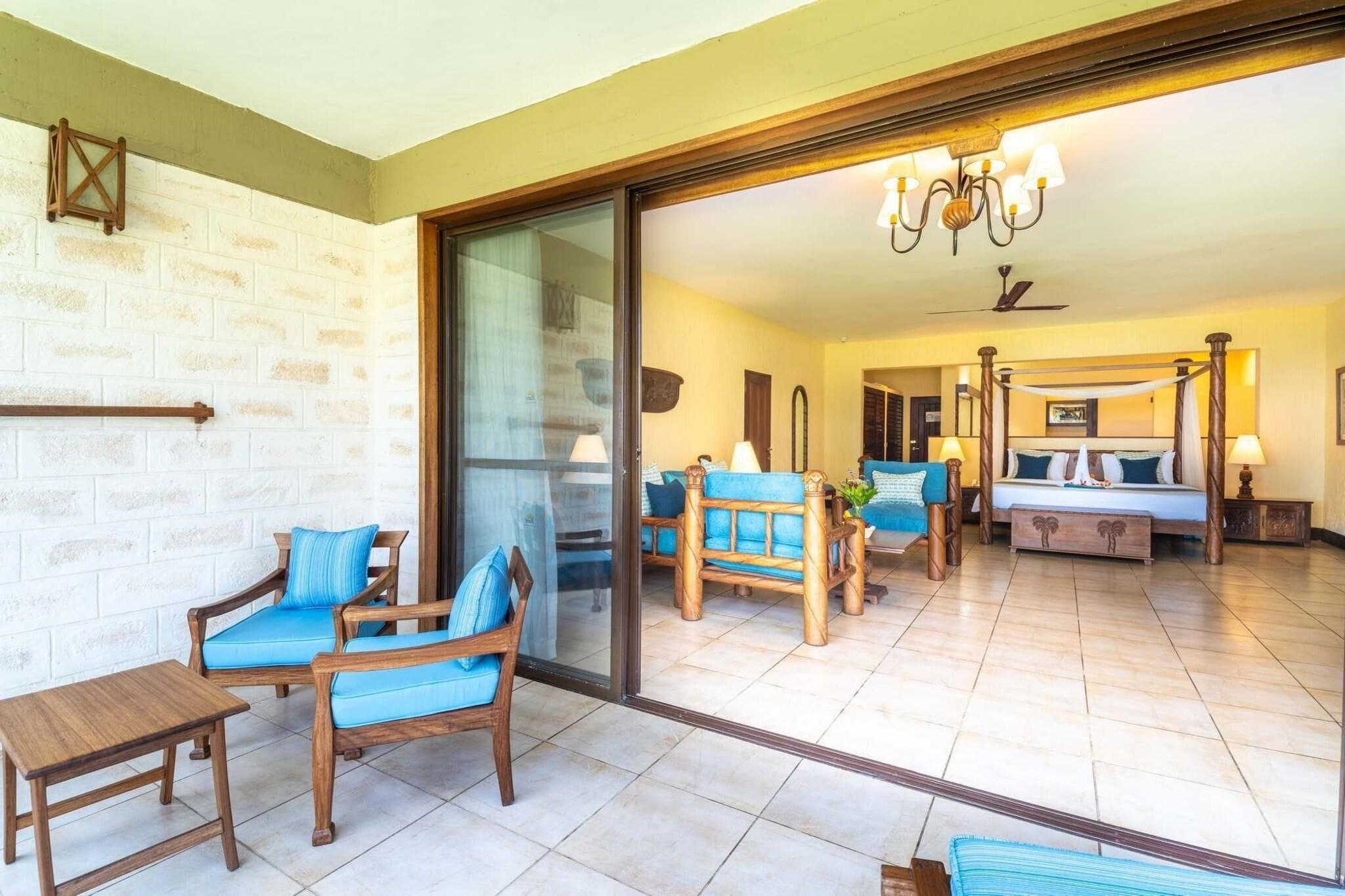 A room with a glass door leading to a balcony, a bed with a canopy, and a seating area with blue cushioned chairs.