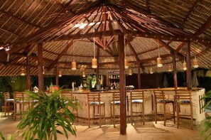 Un área de bar al aire libre con techo de paja, taburetes de madera y mostrador de bar.