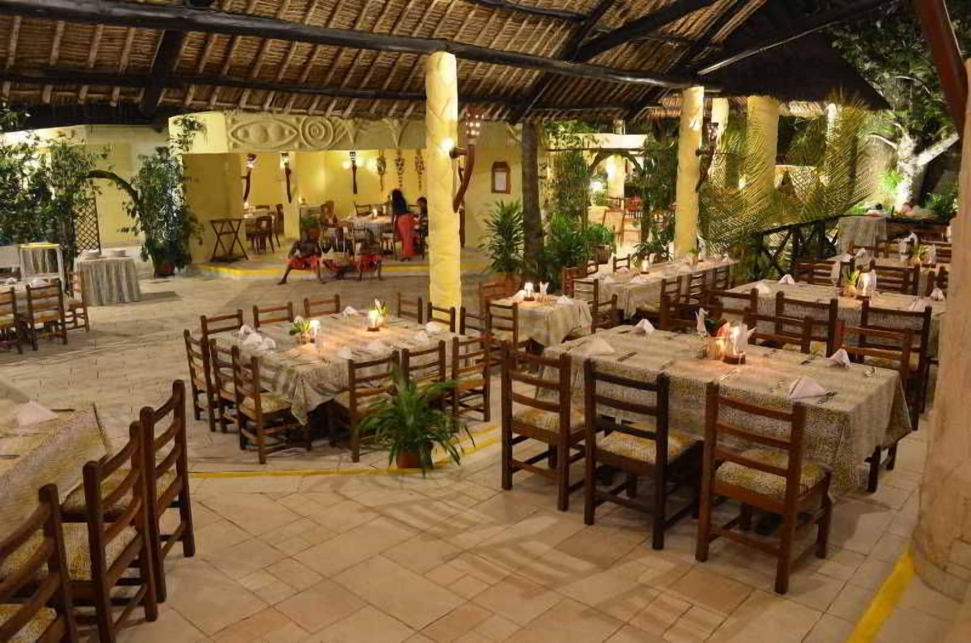 Un restaurante al aire libre con mesas y sillas de madera, rodeado de plantas y un techo de paja.