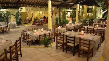 Un restaurante al aire libre con mesas y sillas de madera, rodeado de plantas y un techo de paja.
