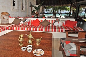 A lounge area with a striped sofa set, a wooden coffee table with traditional tea set, and a view of the beach. - Baobab Beach Resort (Diani Beach)