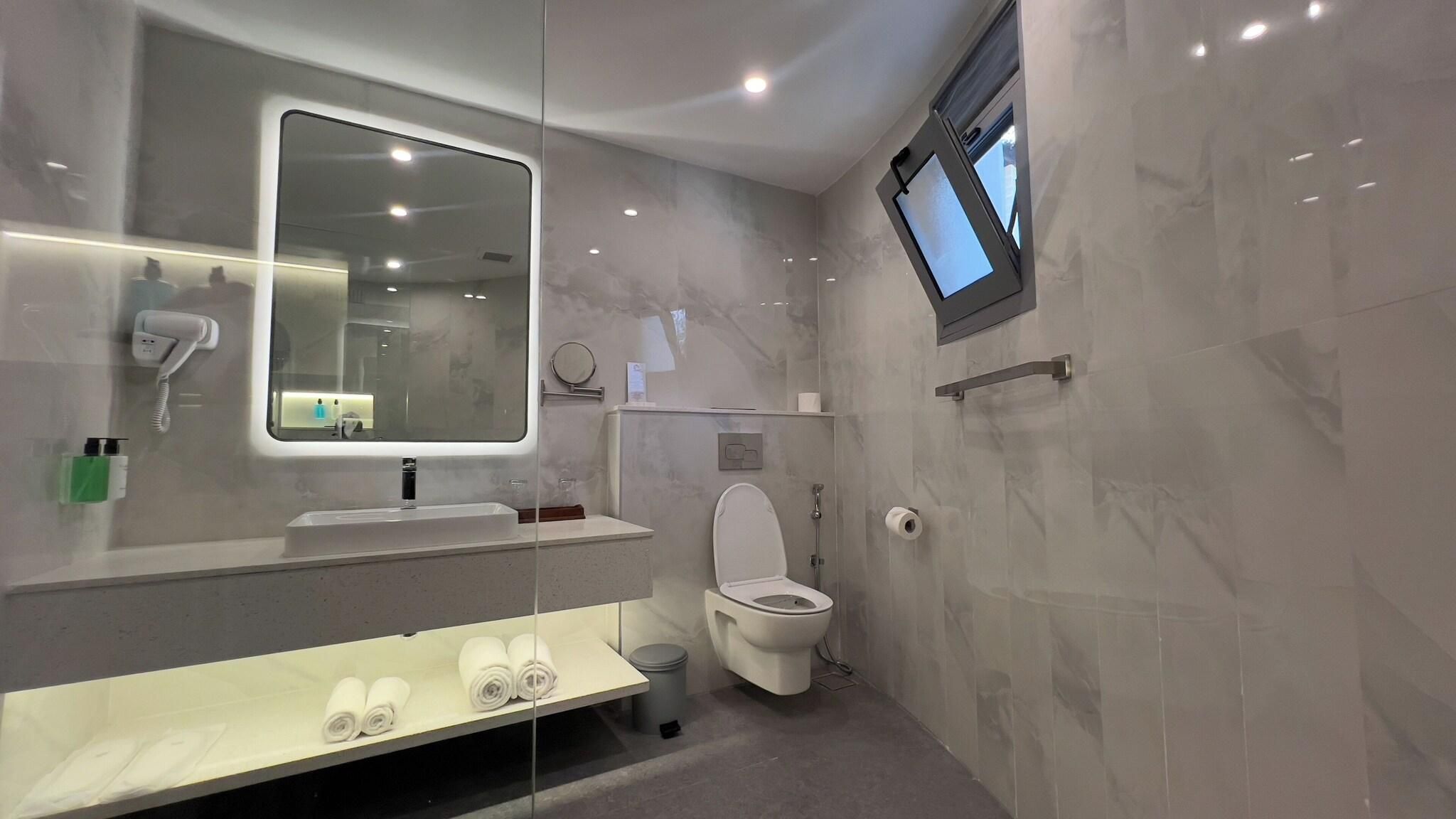 A modern bathroom with a large mirror, a built-in hairdryer, a toilet, and a shelf with towels.