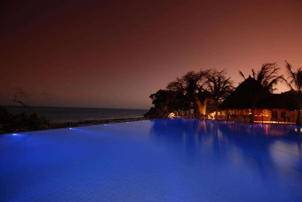 A nighttime view of a pool with a sunset in the background, surrounded by palm trees and a beach.