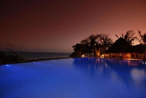 Vista nocturna de una piscina con un atardecer de fondo, rodeada de palmeras y una playa.