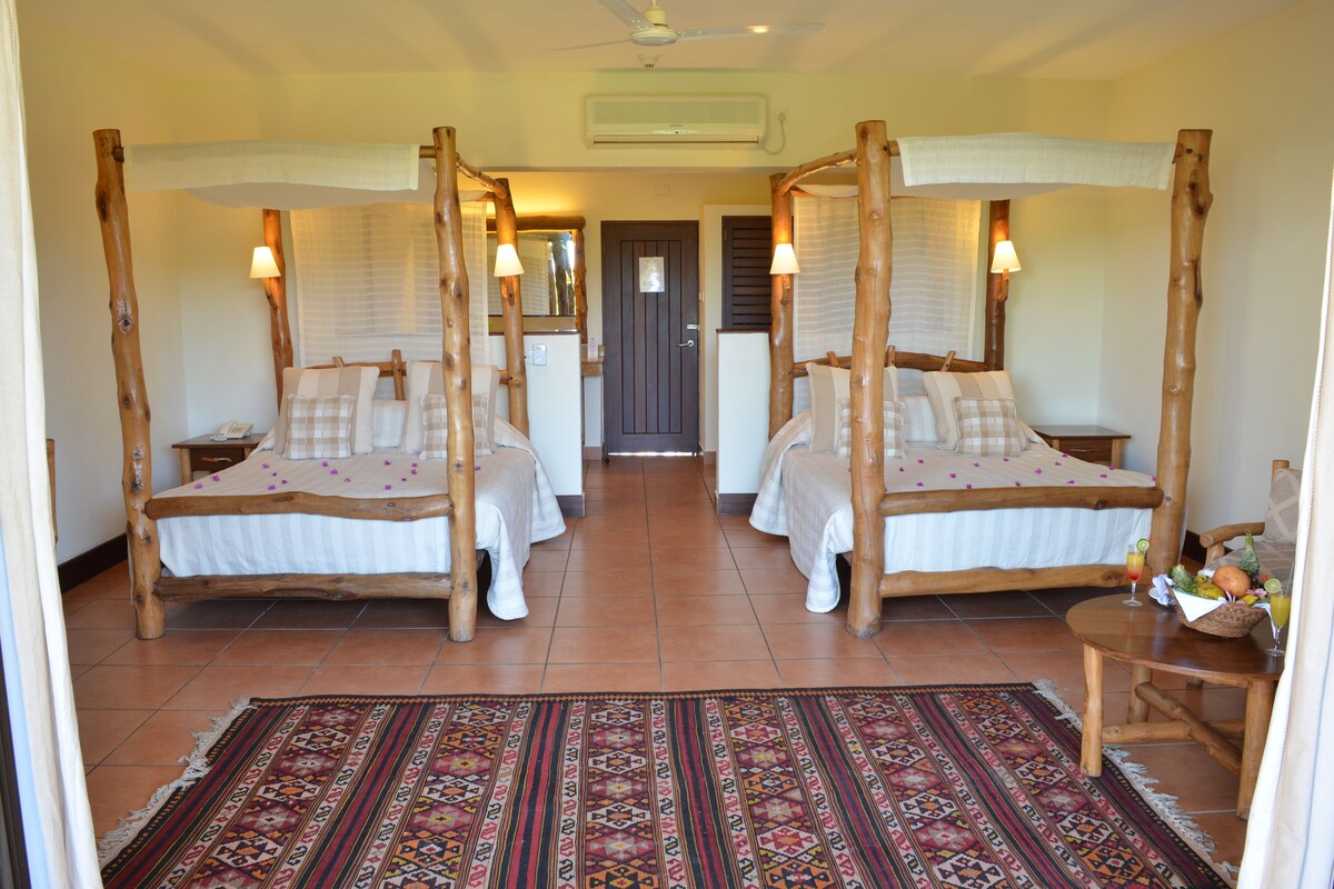 A room with two wooden four-poster beds, a patterned rug, and a small table with a fruit basket and drinks.