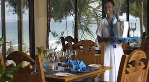 A dining area with wooden furniture, a table set for two, and a view of the ocean through large windows. - Baobab Beach Resort (Diani Beach)