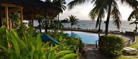 A pool area with a view of the ocean, surrounded by palm trees and a thatched-roof structure.
