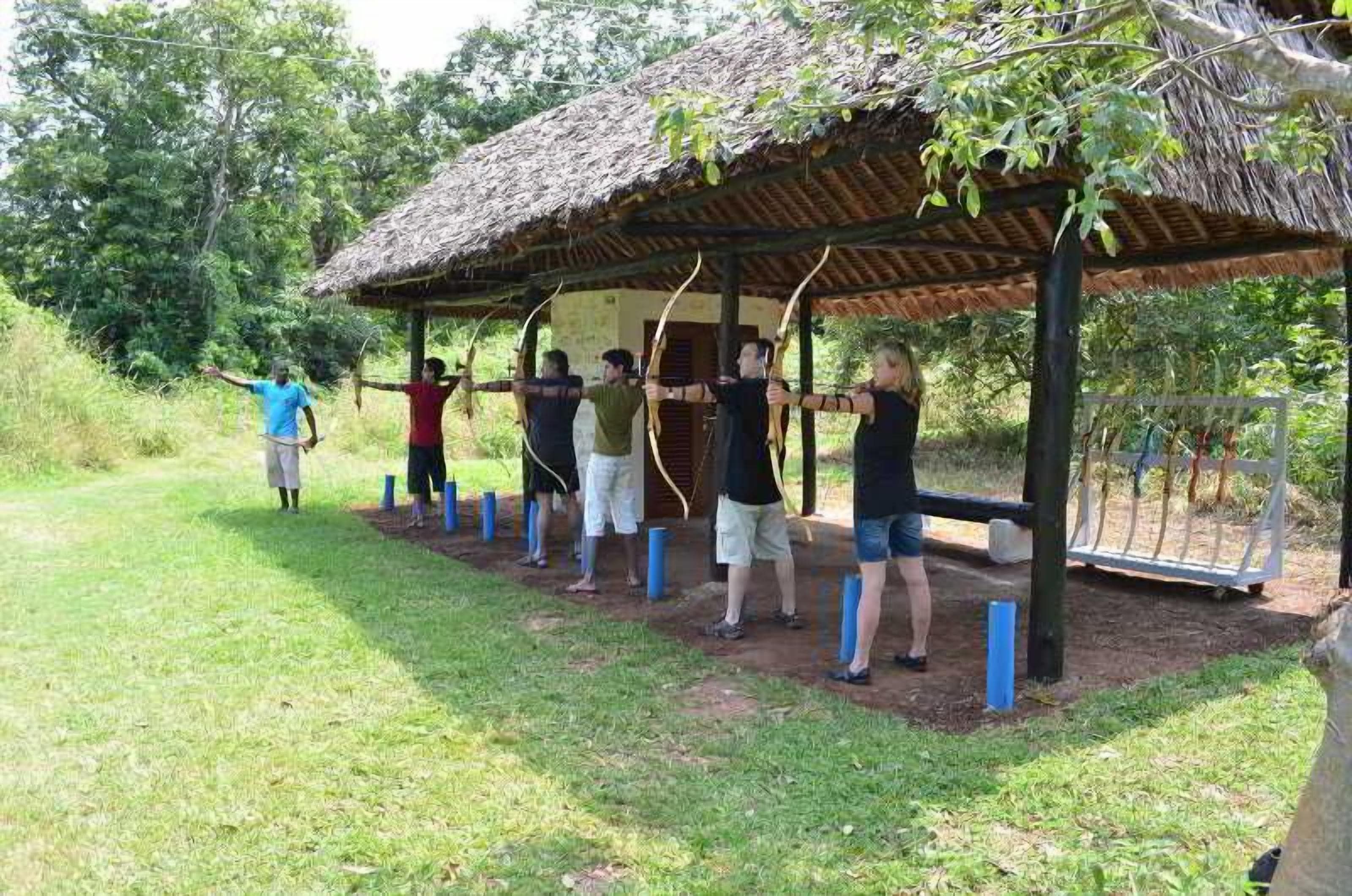 A group of people practicing archery under a thatched-roof pavilion in a grassy outdoor area.