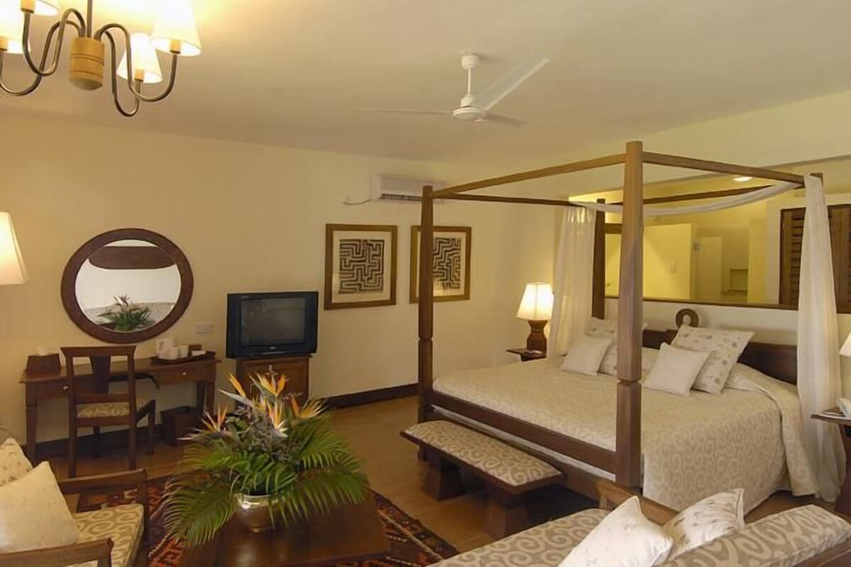 A four-poster bed with a canopy, a television on a stand, a desk with a chair, and a round mirror on the wall.