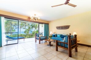 A spacious living room with a large sliding glass door leading to a balcony with a view of palm trees and the ocean. There is a blue sofa, two armchairs, and a wooden coffee table. - Baobab Beach Resort (Diani Beach)