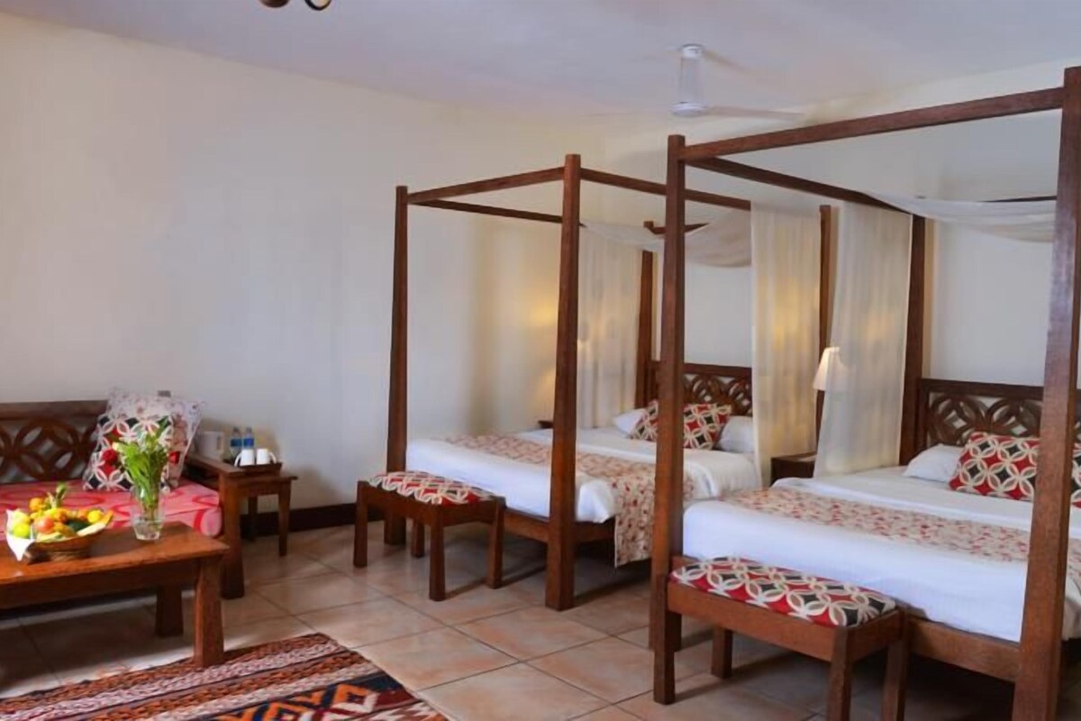 A room with two single beds, a wooden bench, a table with a vase of flowers, and a patterned rug.