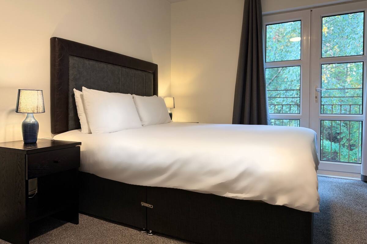 Ein Hotelzimmer mit einem großen Bett, einem Nachttisch mit Lampe und Blick ins Grüne durch das Fenster.