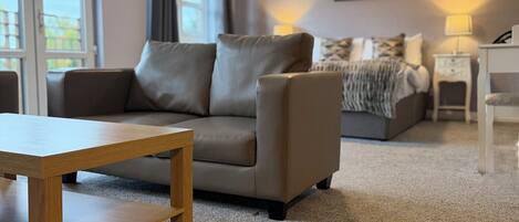 A living room with a sofa, a wooden coffee table, a lamp, and a bed in the background.