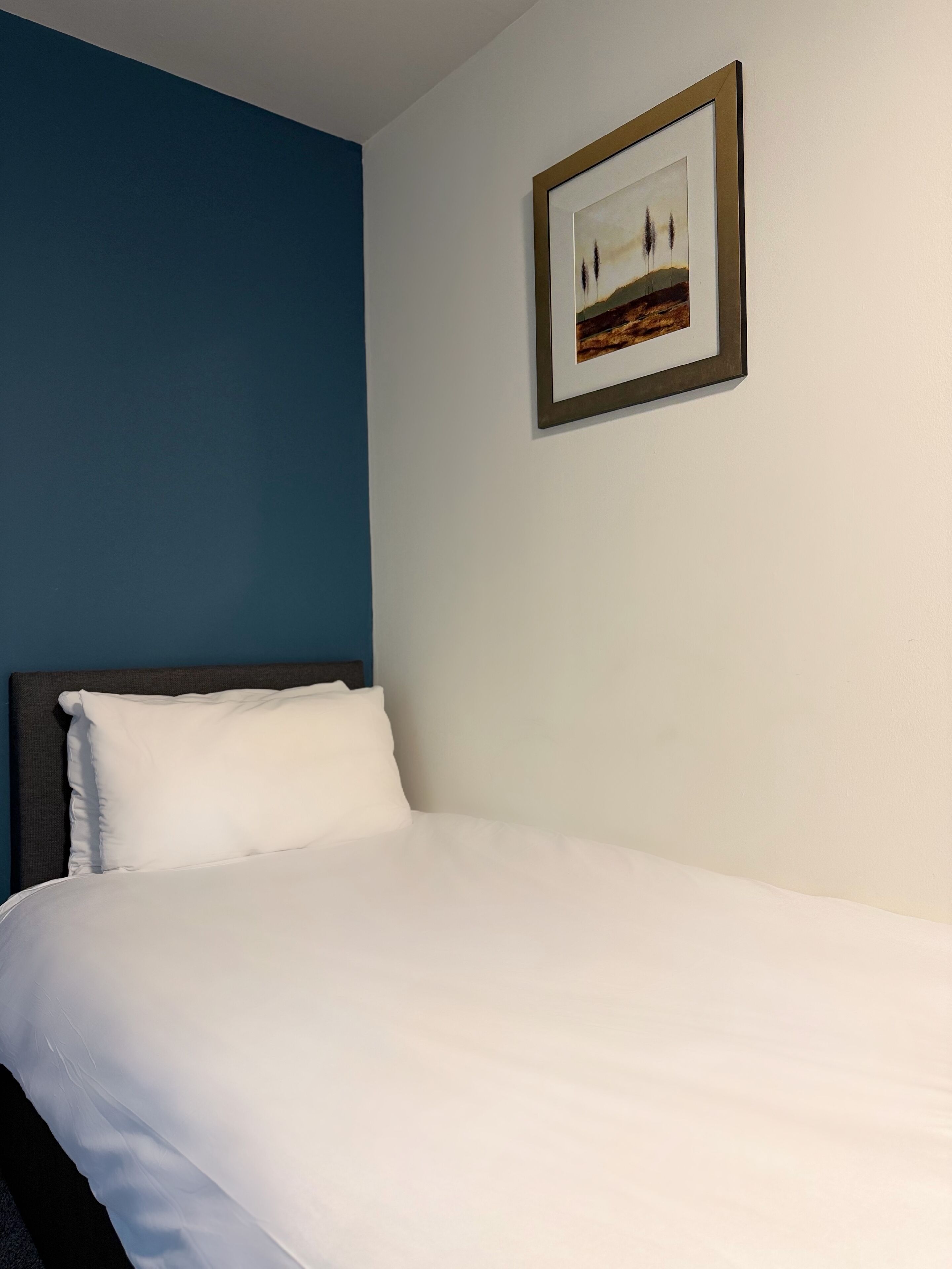 Una cama bien hecha con ropa blanca en una habitación con una pared azul decorativa y un cuadro de paisaje colgado encima.