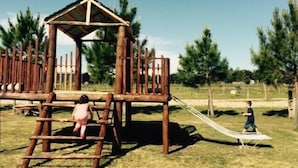 A wooden play structure with a slide and climbing elements in a grassy park area. - Posada de Campo Lo de Fatsia (Colonia del Sacramento)