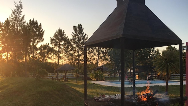 A gazebo with a fire pit in the center, surrounded by a grassy area with trees and a pool in the background. - Posada de Campo Lo de Fatsia (Colonia del Sacramento)