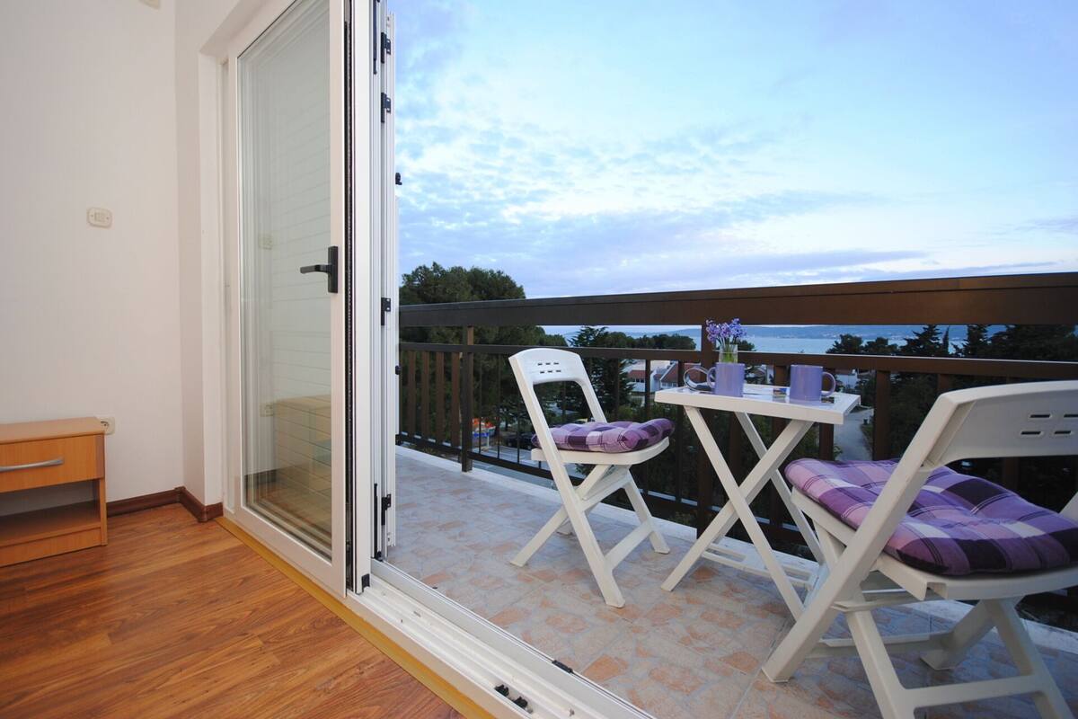 A balcony with a sliding glass door, two white chairs with purple cushions, and a small wooden bench.