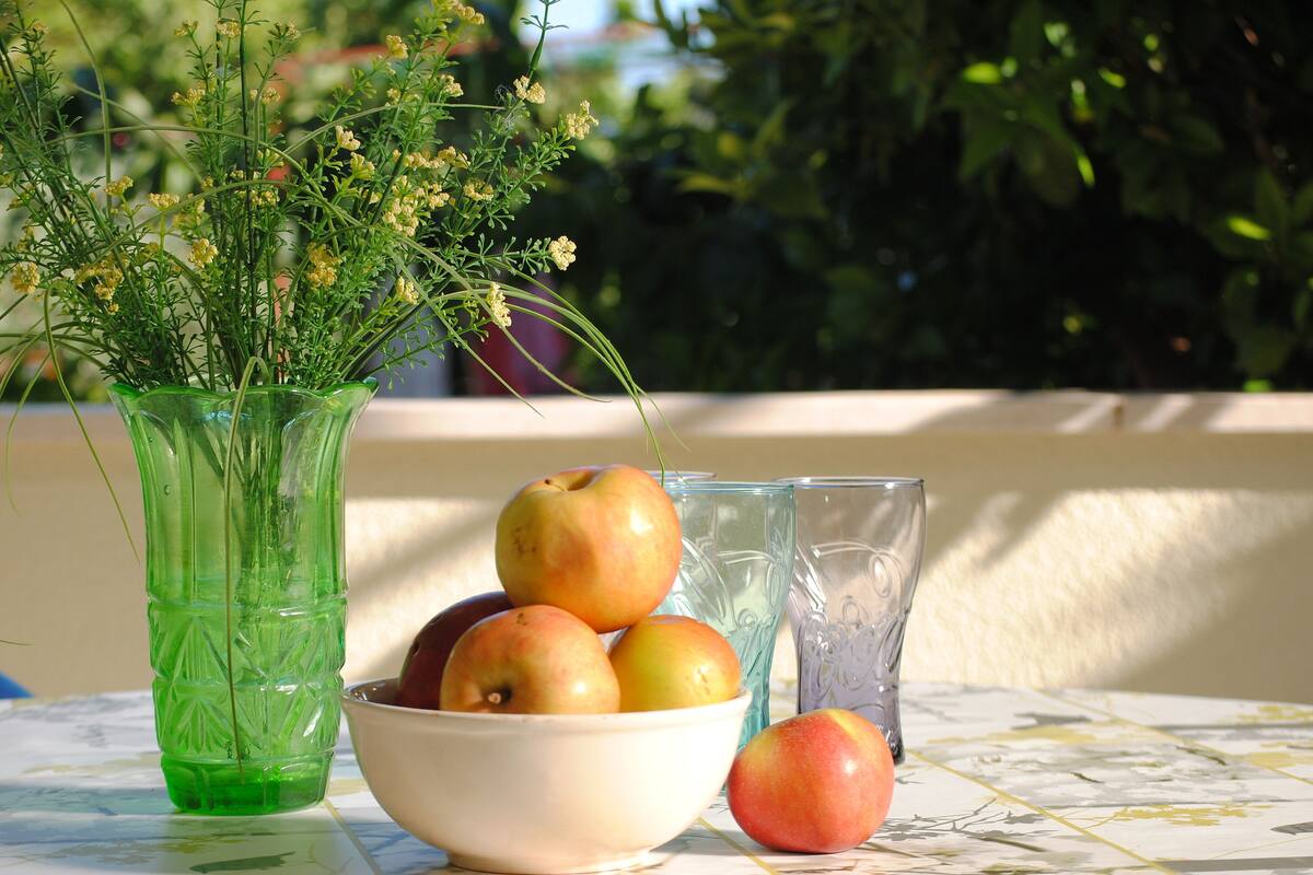 A table with a bowl of apples, a glass, and a green vase with flowers.