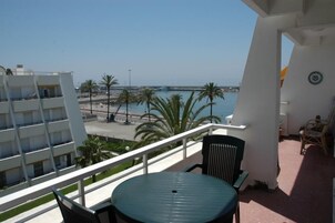 Un balcón con mesa y sillas, con vista a una playa con palmeras y cielo despejado.