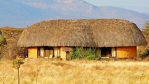 Una cabaña con techo de paja y techo plano, rodeada de pastizales secos y montañas al fondo.