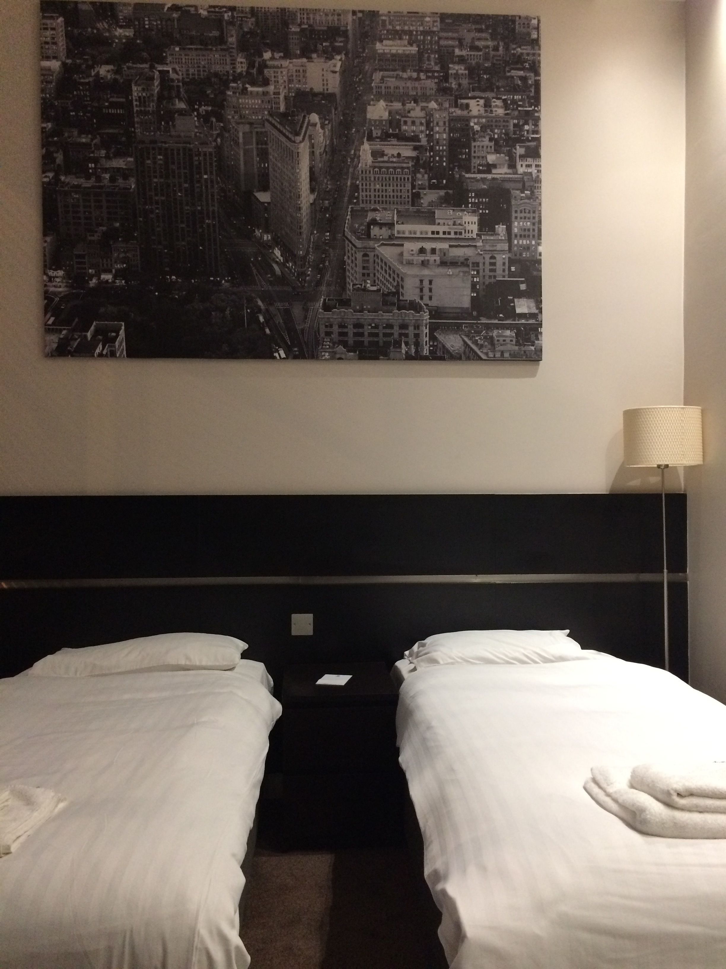 A hotel room with two beds, a cityscape painting on the wall, a bedside lamp, and a dark headboard.