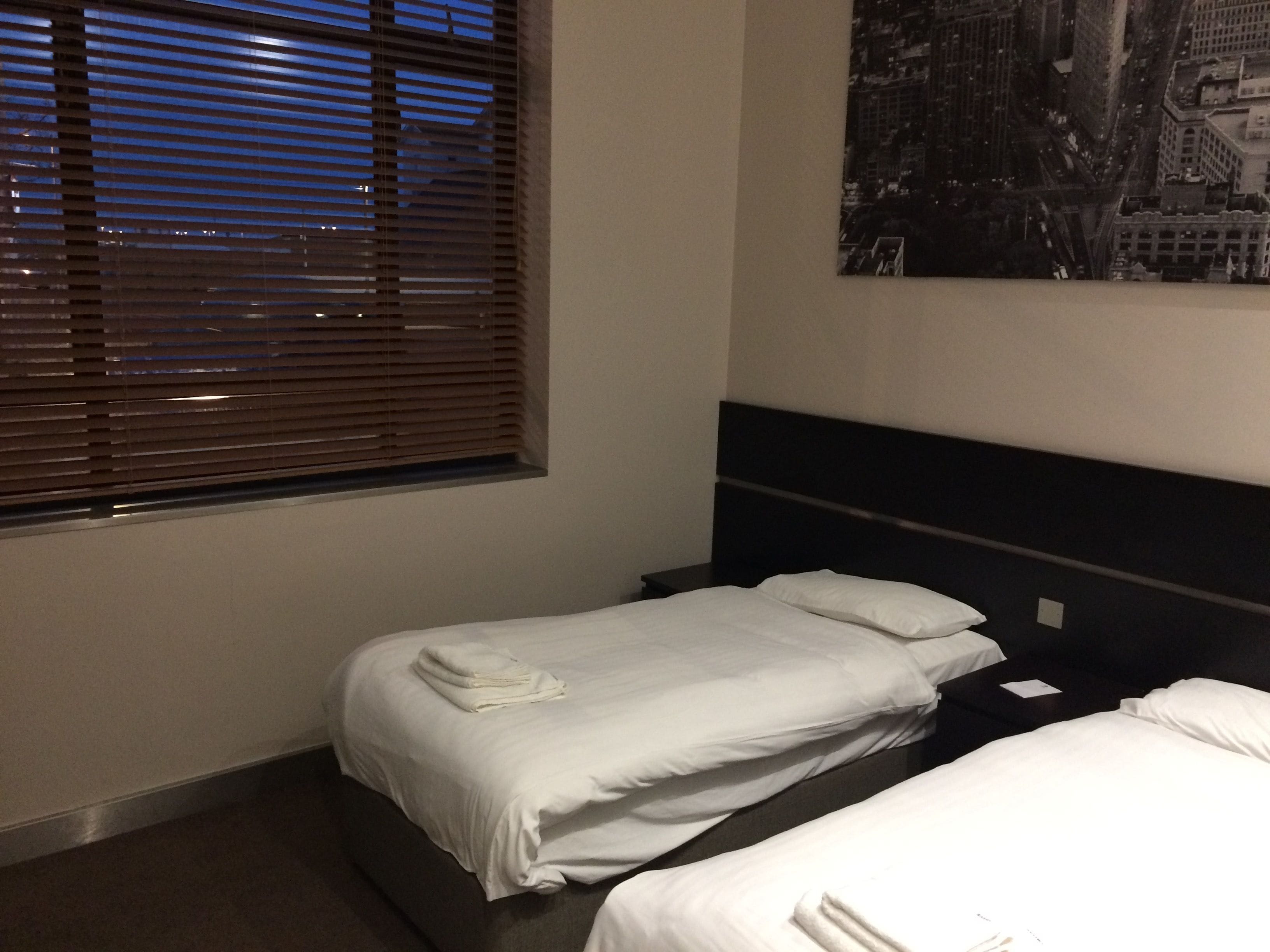 A hotel room with two beds, a window with blinds, a cityscape poster on the wall, and a dark headboard.