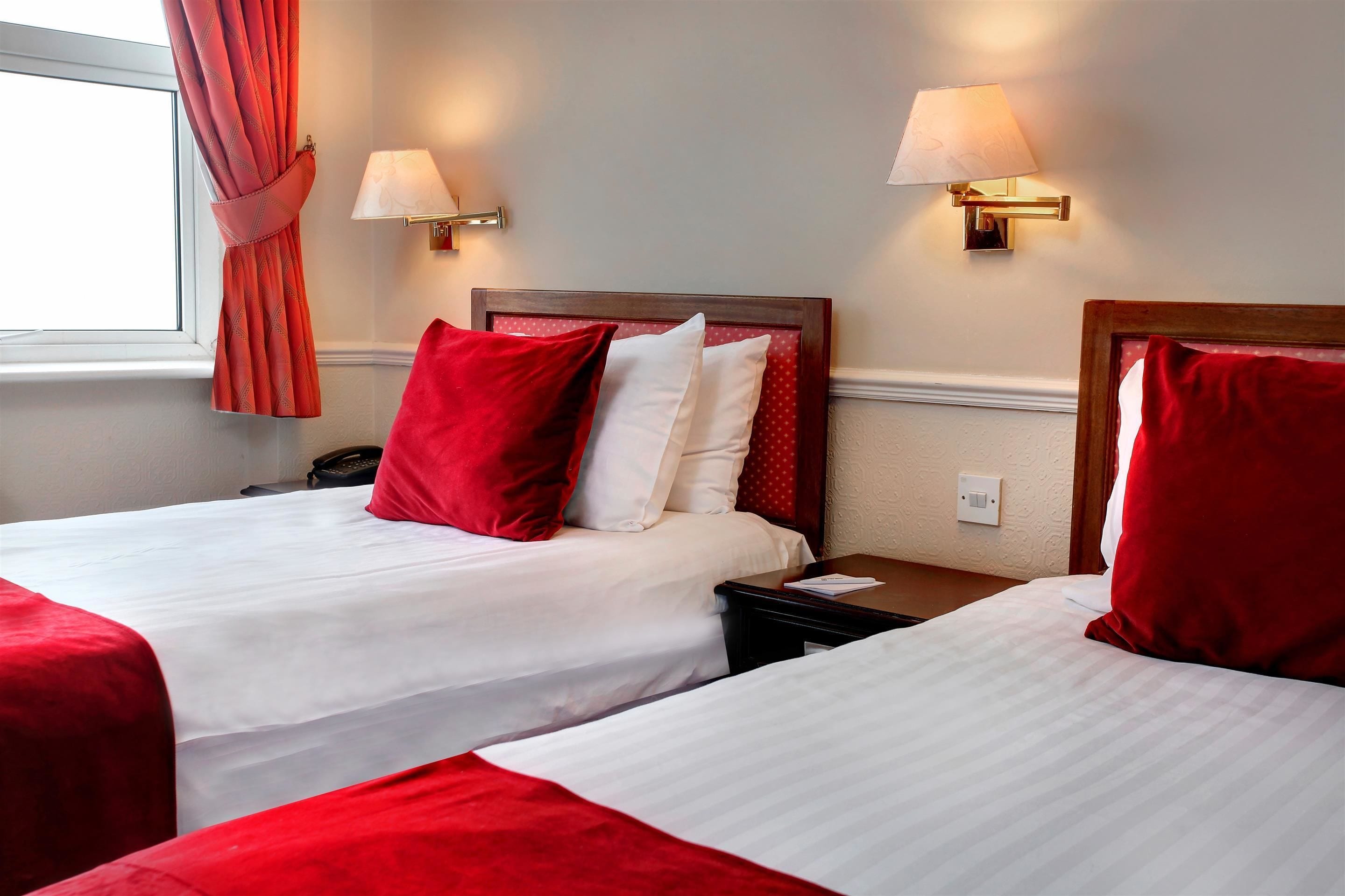 A hotel room with two beds, each with red and white bedding, and two wall-mounted lamps.