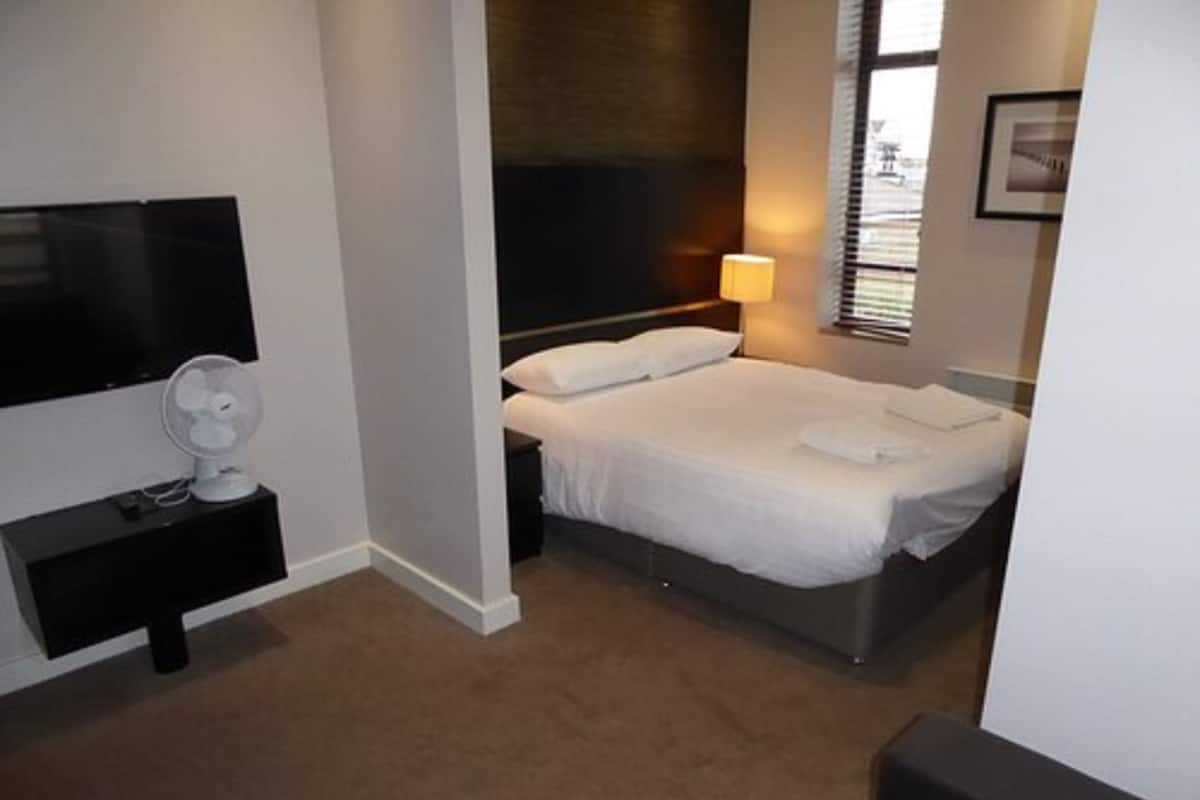 Ein Schlafzimmer mit Bett, Fernseher, Nachttisch mit Lampe und einem Fenster mit Jalousien.