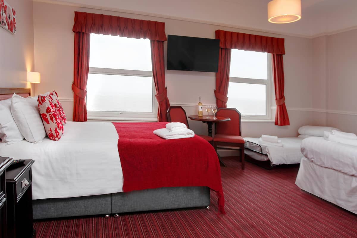 Ein Hotelzimmer mit einem Bett, einem Schreibtisch mit Stuhl, einem kleinen Tisch mit Flasche und Glas sowie zwei Fenstern mit Vorhängen.