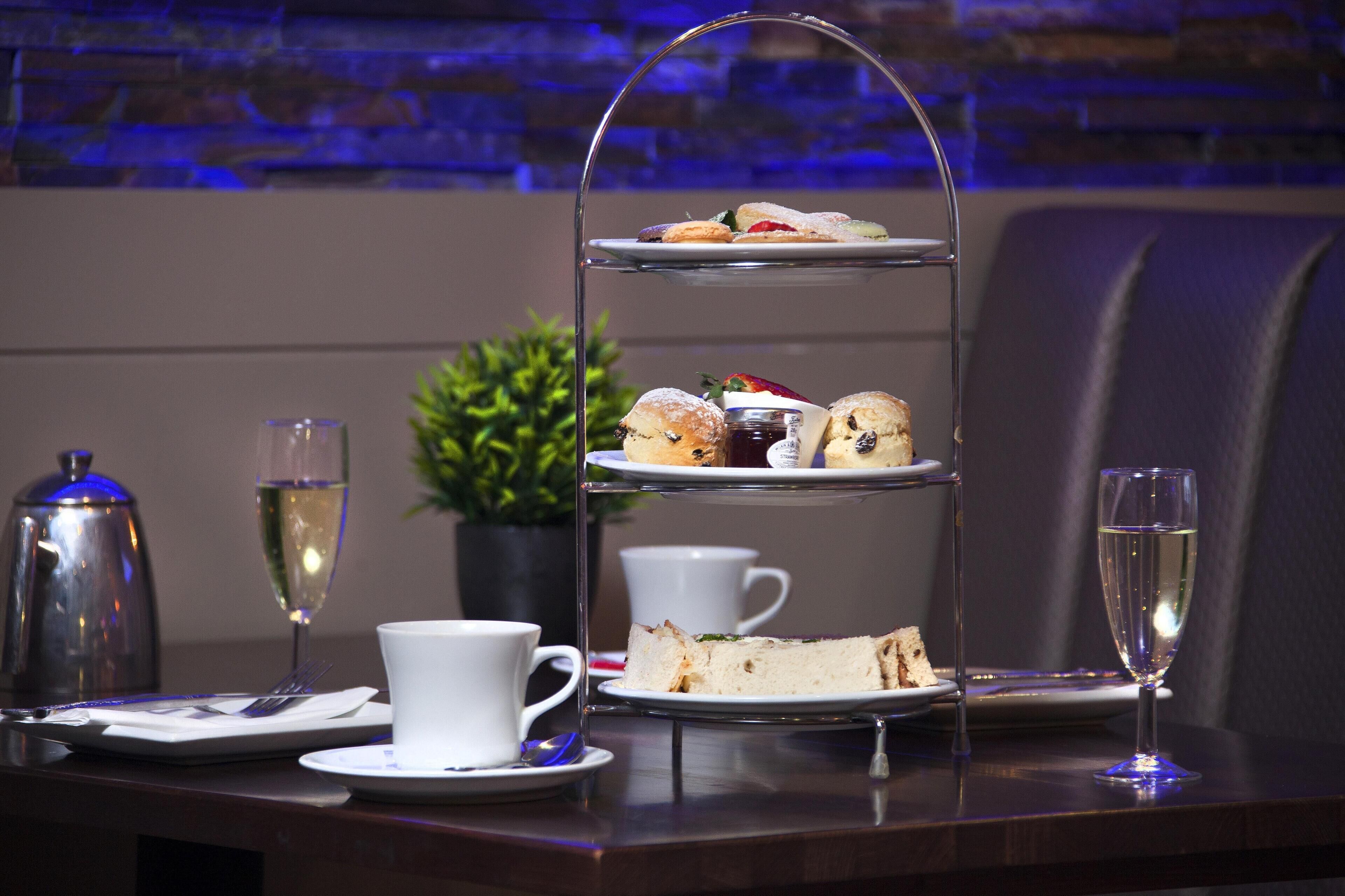Ein dreistöckiges Kuchenbuffet mit Sandwiches und Gebäck, einer Kaffeetasse, einem Glas Champagner und einem Teekännchen auf einem Tisch.