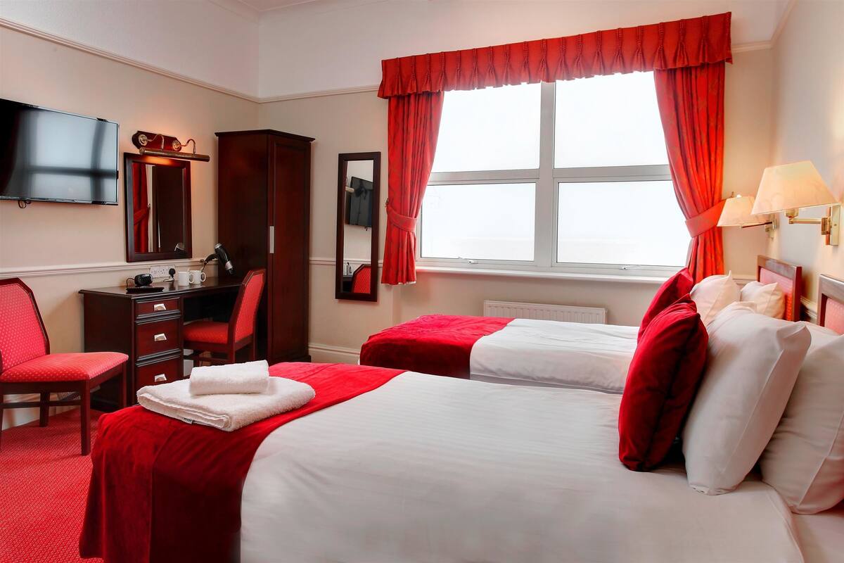 Ein Hotelzimmer mit zwei Betten, einem Schreibtisch mit Stuhl, einem Fernseher und einem Fenster mit Vorhängen.
