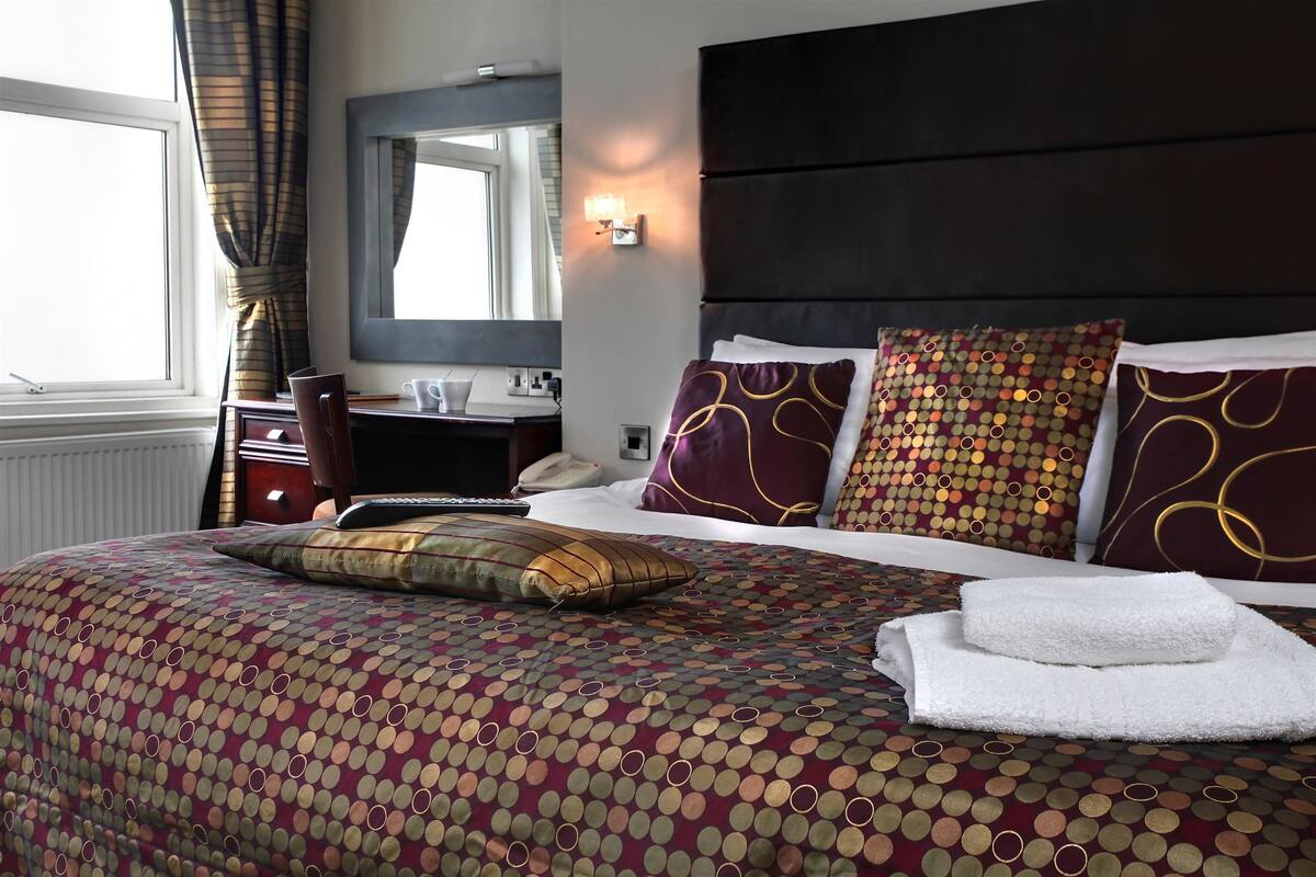 Ein Hotelzimmer mit einem Bett, einem Nachttisch, einer Lampe, einem Spiegel und einem Fenster mit Vorhängen.