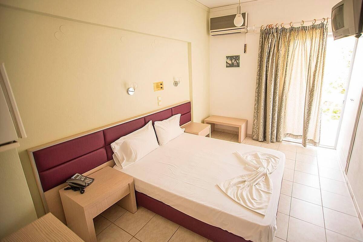 Ein Hotelzimmer mit einem großen Bett, einem Nachttisch, einem kleinen Schreibtisch, einer Klimaanlage, einem Fernseher und einem Fenster mit Vorhängen.