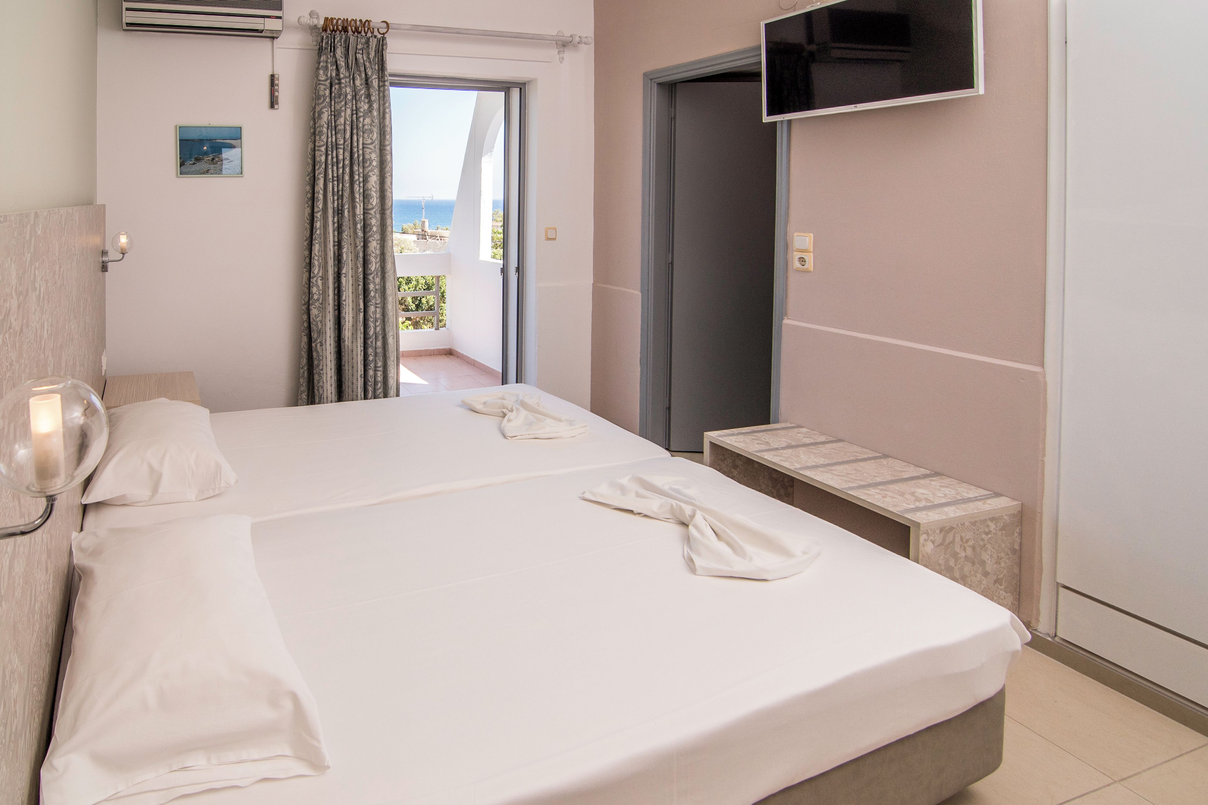 A hotel room with a bed, a TV mounted on the wall, and a view of the sea through an open door.