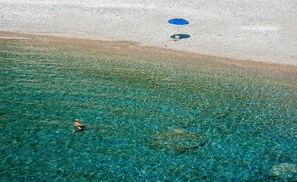 A clear view of a beach with a person swimming in the water and another person sitting under a blue umbrella on the sand. - Megim (Kantanos-Selino)