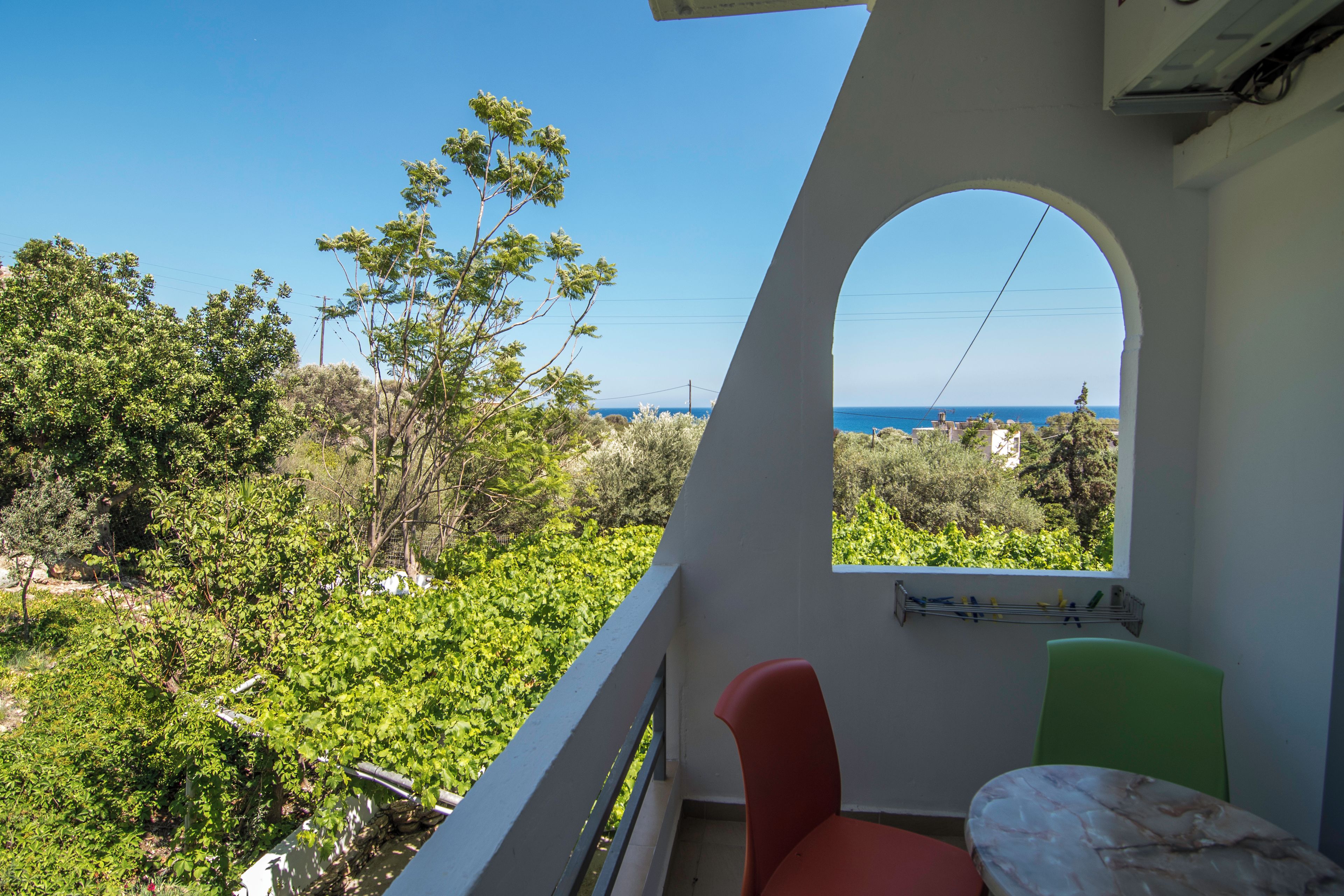 A balcony with a view of the sea and greenery, a red chair, and a green chair.
