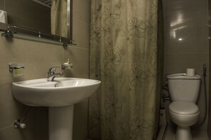 Kylpyhuoneessa on valkoinen pesuallas, sen yläpuolella peili, wc-istuin ja kukkakuviollinen suihkuverho.