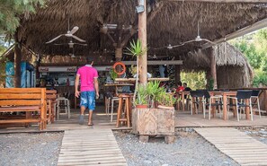 An outdoor seating area with wooden tables and chairs, a thatched roof, and a Coca-Cola refrigerator. - Megim (Kantanos-Selino)