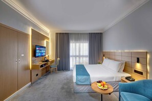 A hotel room with a bed, a desk with a computer, a TV, a chair, a small table with fruit, and a sofa. - Lotus Grand Hotel Dubai (Dubai)