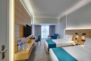 A hotel room with two beds, a desk with a flat-screen TV, a chair, and a small table with water bottles and fruit.