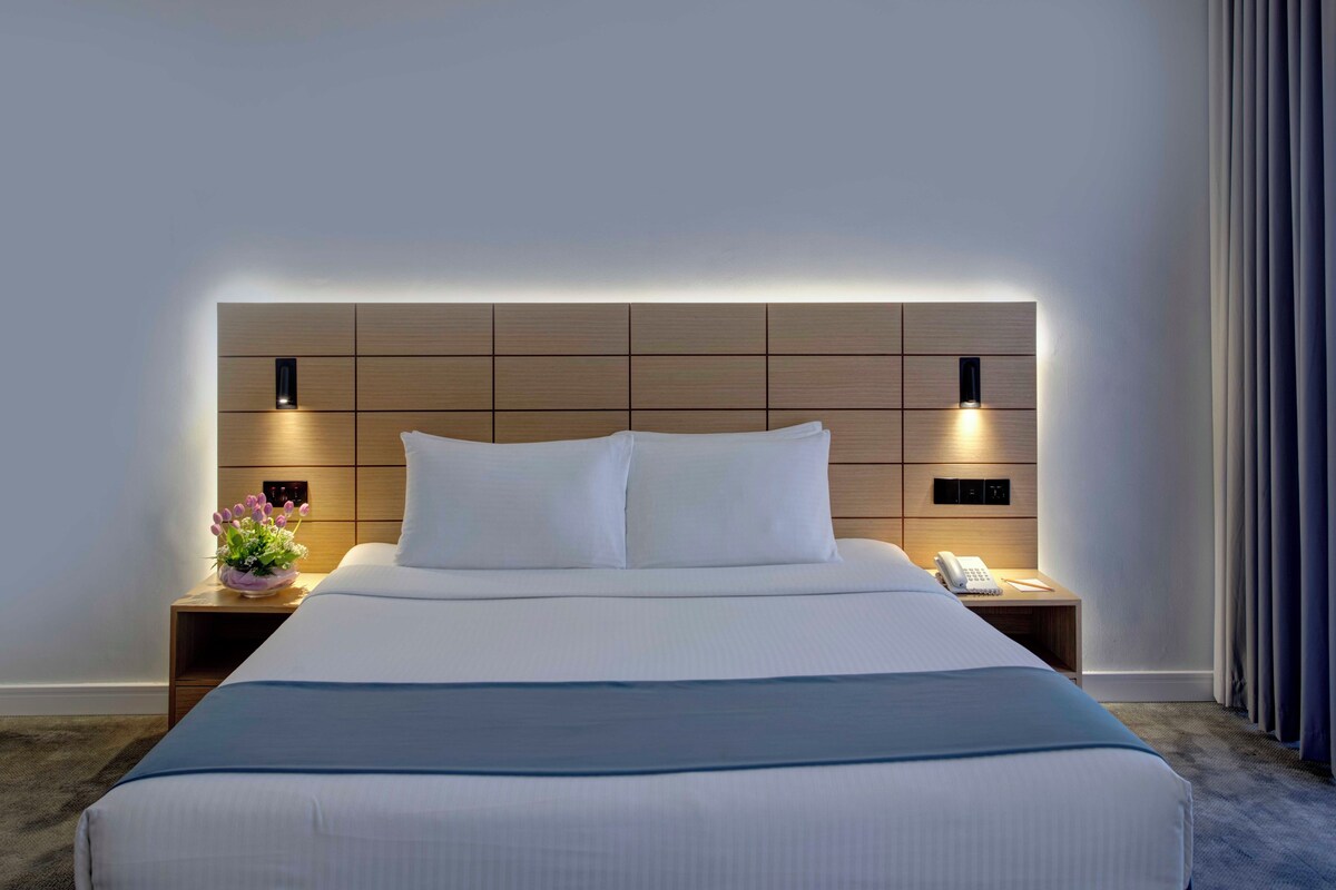 A modern hotel room with a large bed, a wooden headboard, bedside tables with lamps, and a small table with a vase of flowers.