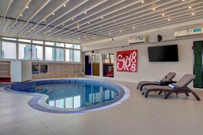 A modern indoor pool area with a curved pool, lounge chairs, a large TV, and a red artwork on the wall. - Lotus Grand Hotel Dubai (Dubai)