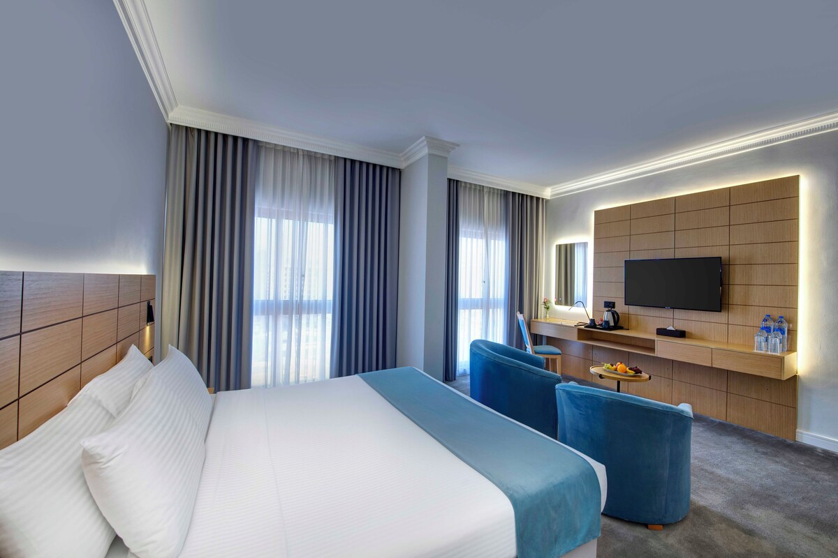 A modern hotel room with a large bed, a desk with a flat-screen TV, and a seating area with a blue chair.