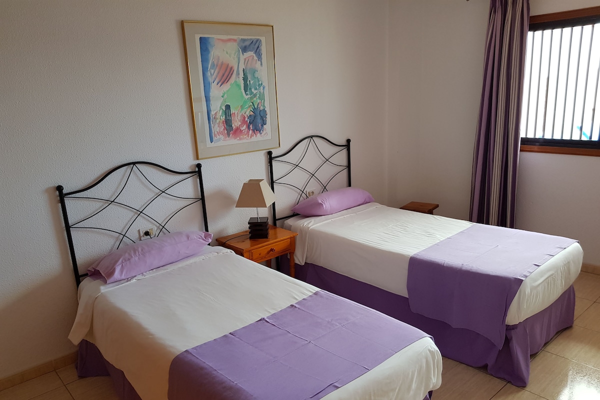 A room with two beds, each with a purple bedspread and white sheets, a small wooden nightstand with a lamp, and a framed abstract painting on the wall.