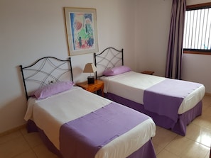 A room with two beds, each with a purple bedspread and white sheets, a small wooden nightstand with a lamp, and a framed abstract painting on the wall. - Las Fuentes (Arona)