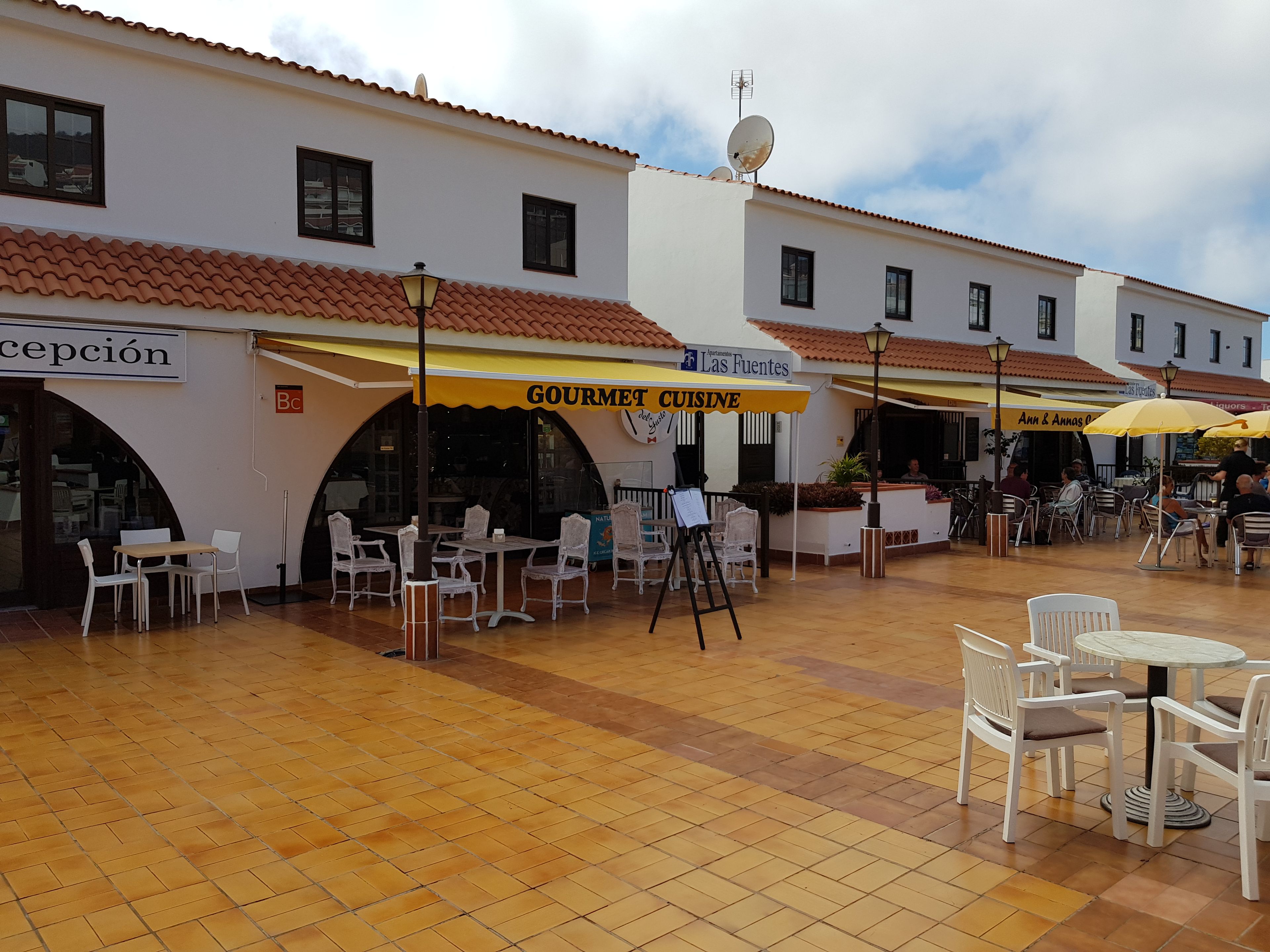 An outdoor cafe area with tables and chairs, a sign for 'Gourmet Cuisine', and a building with a tiled roof and arches.