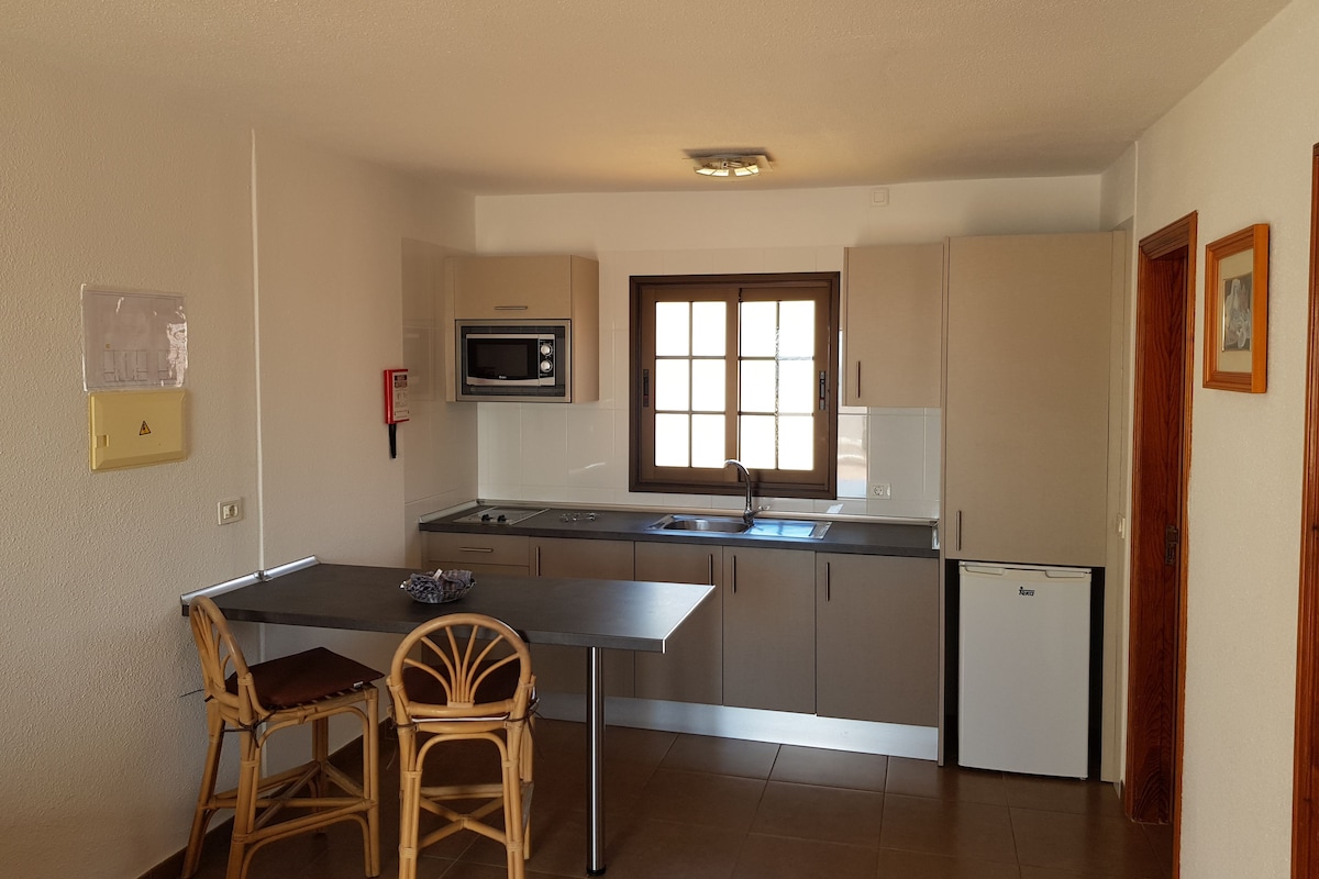 A compact kitchen with a microwave, sink, and small refrigerator. There are two wooden chairs at a table, and a window above the sink.