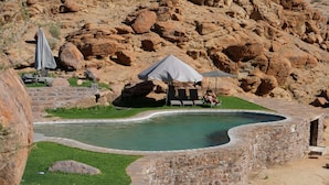 A swimming pool with a stone wall, lounge chairs, and umbrellas. - Canyon Lodge (Fish River Canyon)