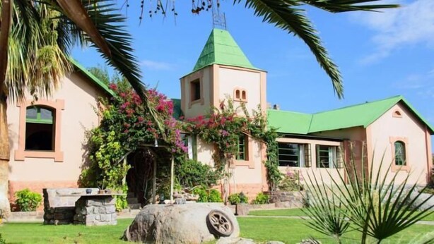 A building with a green roof, a tower, and a garden with pink flowers. - Canyon Lodge (Fish River Canyon)